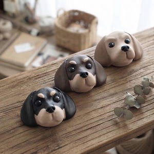 May include: Three sculpted dog head figurines arranged in a row. The first is black and tan, the second is dark brown and tan, and the third is light brown. Each figurine features rounded ears, black eyes, and a black nose, crafted with smooth surfaces.