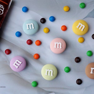 Könnte beinhalten: Eine Sammlung runder, bunter, bonbonartiger Dekorationen mit dem Buchstaben "M" in Weiß auf jeder. Die Dekorationen sind auf einer weißen Oberfläche angeordnet, mit kleinen, bunten Bonbons, die um sie herum verstreut sind.