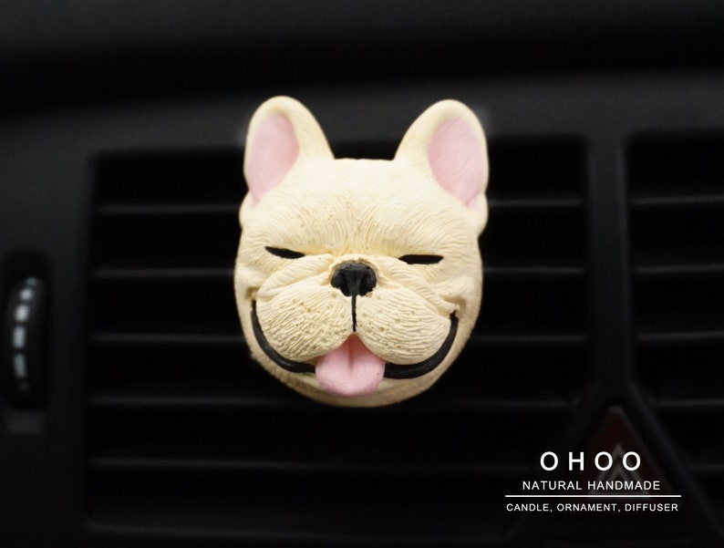 May include: A cream-colored French bulldog head ornament with pink inner ears and tongue, black nose and mouth, and closed eyes. The text OHOO NATURAL HANDMADE CANDLE, ORNAMENT, DIFFUSER is printed in white in the bottom right corner of the image.