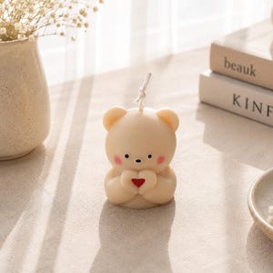 K&ouml;nnte beinhalten: Eine beigefarbene, b&auml;renf&ouml;rmige Kerze mit einem kleinen wei&szlig;en Docht an der Oberseite, schwarzen Augen, einer schwarzen Nase und rosa Wangen. Der B&auml;r h&auml;lt ein kleines, rotes Herz zwischen seinen Pfoten. Die Kerze hat eine glatte, matte Oberfl&auml;che.
