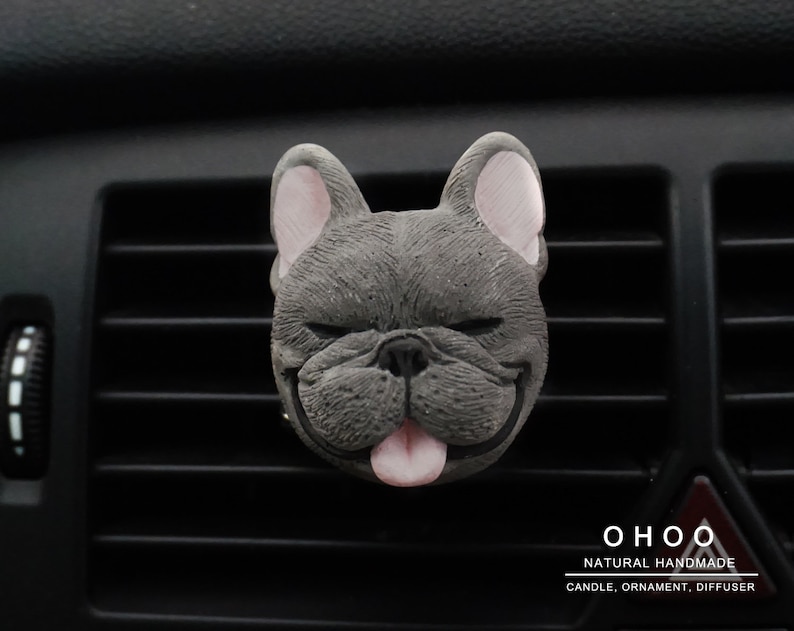 May include: A gray French bulldog head ornament with closed eyes and a pink tongue sticking out. The text OHOO NATURAL HANDMADE CANDLE, ORNAMENT, DIFFUSER is printed in white in the bottom right corner of the image.