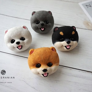 May include: Four small ceramic figurines of Pomeranian dogs in different colours: white, grey, black, and orange. Each figurine has a cute, cartoon-like expression with a pink tongue sticking out.