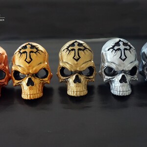 May include: Five skull ornaments arranged in a row, each featuring a cross symbol on the forehead. The skulls are finished in metallic copper, bright gold, muted gold, silver, and dark grey. Text in the top left corner reads OHOO NATURAL HANDMADE CANDLE, ORNAMENT, DIFFUSER.