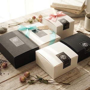 May include: A collection of rectangular and square gift boxes in matte black and white finishes. Each box features a black rectangular label with a white floral graphic and the text ONOO. Some boxes are adorned with light blue or pink satin ribbons.