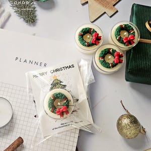 May include: Christmas candles with a wreath design, in clear containers. The wreaths are green with red bows and gold details. The image includes a card that says "Merry Christmas". The candles are handmade.