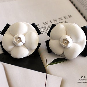 Puede incluir: Dos adornos de flores de camelia blancas con pétalos en capas y una pequeña perla central. Cada flor está montada sobre un lazo de cinta negra. El texto OHOO NATURAL HANDMADE CANDLE, ORNAMENT, DIFFUSER está impreso en la esquina inferior derecha.