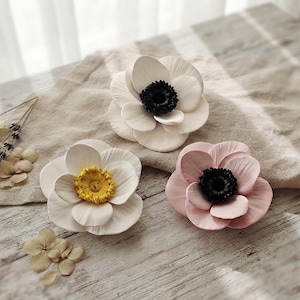 Puede incluir: Tres flores decorativas hechas a mano con pétalos en capas. Una flor es blanca con centro negro, otra es blanca con centro amarillo y otra es rosa claro con centro negro. Las flores están dispuestas sobre una superficie texturizada.