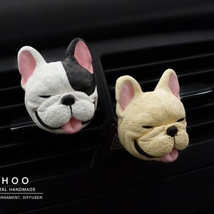 May include: Two French bulldog head-shaped car air vent clips. One is white with a black patch over one eye and ear, the other is cream-colored. Both have pink ears, black noses, and tongues sticking out. Text reads OHOO NATURAL HANDMADE CANDLE, ORNAMENT, DIFFUSER.