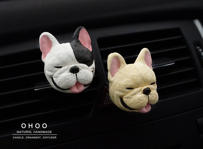 May include: Two French bulldog head-shaped car air vent clips. One is white with a black patch over one eye and ear, the other is cream-colored. Both have pink ears, black noses, and tongues sticking out. Text reads OHOO NATURAL HANDMADE CANDLE, ORNAMENT, DIFFUSER.