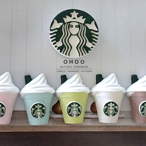 Könnte beinhalten: Fünf bunte, handgefertigte Kerzenornamente in Form von Starbucks Frappuccinos, jedes mit einer weißen Schlagsahne und einem grünen Strohhalm. Das Starbucks-Logo befindet sich auf jeder Tasse. Ein großes Starbucks-Logo und der Text "OHOO NATURAL HANDMADE" befinden sich darüber.