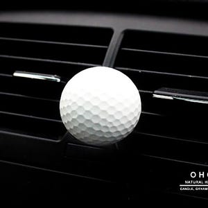 Könnte beinhalten: Weißer Golfball-Lufterfrischer für das Auto, der an einer Lüftungsschlitz im Auto hängt. Der Lufterfrischer ist ein weißer Golfball mit Dellen. Der Lüftungsschlitz ist aus schwarzem Kunststoff.