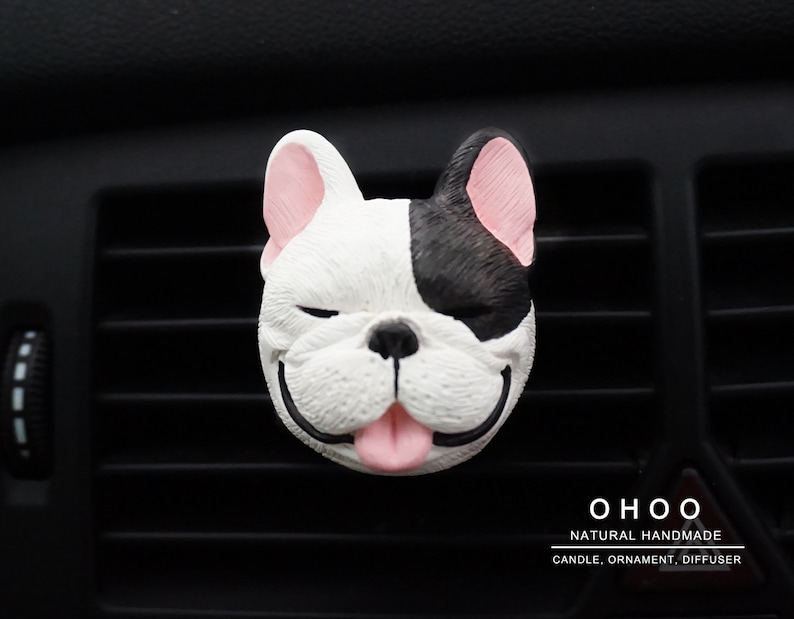 May include: A French bulldog head car air freshener ornament featuring a black and white face with pink ears and tongue, closed eyes, and a smiling expression. The text OHOO NATURAL HANDMADE CANDLE, ORNAMENT, DIFFUSER is printed in the bottom right corner.