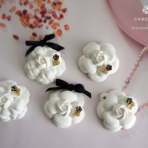 May include: Five white ceramic camellias with black bows and gold and black embellishments. The camellias are arranged on a light pink surface. The text "CAMELLIA DESIGNED BY OHOO" is visible in the top right corner of the image.