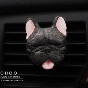 May include: A black French bulldog head ornament with pink inner ears and a pink tongue sticking out. The text O H O O NATURAL HANDMADE CANDLE, ORNAMENT, DIFFUSER is printed in white below the dog figure on the black car air vent slats.