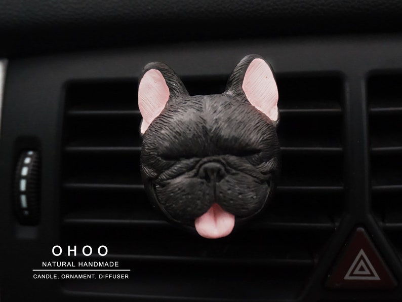 May include: A black French bulldog head ornament with pink inner ears and a pink tongue sticking out. The text O H O O NATURAL HANDMADE CANDLE, ORNAMENT, DIFFUSER is printed in white below the dog figure on the black car air vent slats.