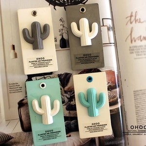 Könnte beinhalten: Vier weiße rechteckige Gips-Lufterfrischer mit Kaktus-Design. Jeder Lufterfrischer trägt den Text "OHOO PLASTER AIR FRESHENER MADE OF PREMIUM PLASTER WITH FRAGRANCE OILS www.ohoo.com.au" aufgedruckt. Die Kakteen sind in verschiedenen Farben: grau, weiß, hellblau und grün.