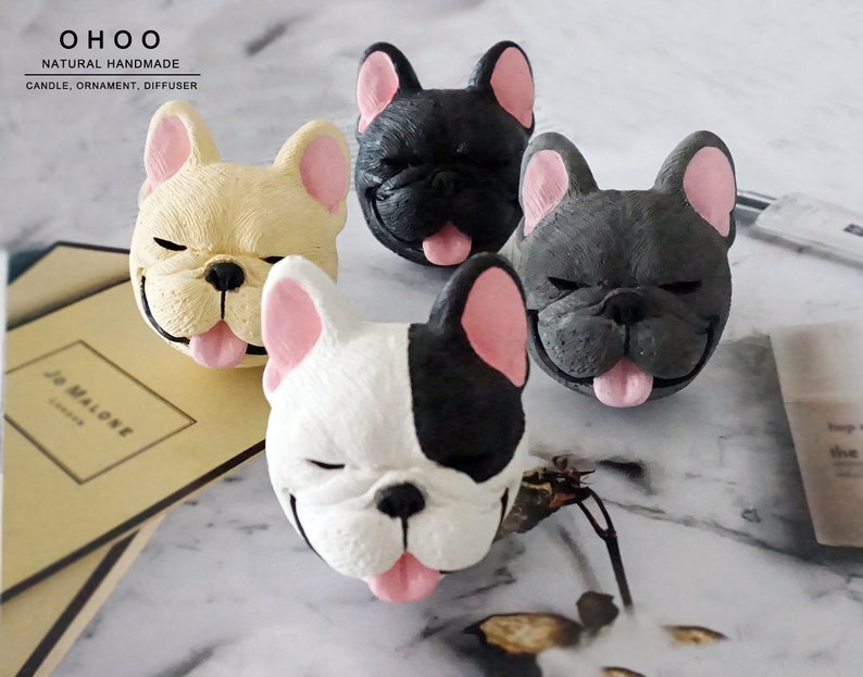 May include: Four French bulldog head ornaments in cream, black, dark gray, and a black and white spotted pattern. Each figure features closed eyes, a black nose, and a pink tongue sticking out. The text OHOO NATURAL HANDMADE CANDLE, ORNAMENT, DIFFUSER is visible.