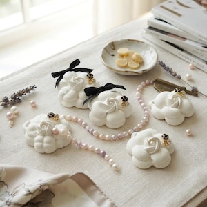 May include: Four white camellia-shaped decorative objects featuring black ribbon bows, small pearl accents, and tiny dark floral embellishments. A long strand of iridescent pink and white pearls is draped among the white flower ornaments.