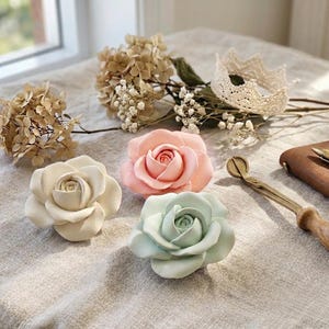 May include: Three ceramic rose sculptures in cream, soft pink, and pale mint green. The roses are arranged alongside dried hydrangea flowers, a small white crocheted crown, a brass tracing wheel with a wooden handle, and a brown leather notebook.