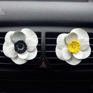 Puede incluir: Dos ambientadores de coche con forma de flor blanca. Uno tiene un centro negro y el otro un centro amarillo. Las flores están sujetas a las rejillas de ventilación de un coche.