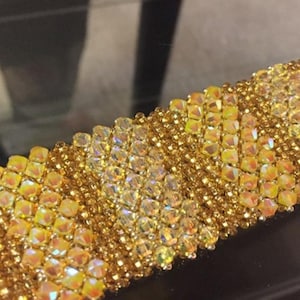 Pode incluir: Close-up de uma pulseira de contas douradas. A pulseira apresenta fileiras de pequenas contas quadradas e iridescentes e contas douradas menores. O design é intrincado e detalhado, com um efeito brilhante. A pulseira parece ser feita à mão.