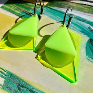 May include: A pair of neon yellow earrings featuring a silver-toned hoop attached to a layered geometric design. Each earring consists of a translucent triangular piece overlapping a solid circular disc, which is set against a larger triangular base.