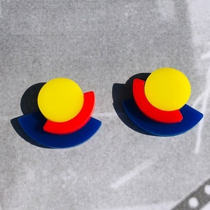 May include: A pair of colorful earrings with a yellow circle, red crescent, and blue half-circle design.