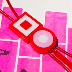 May include: A red and white geometric pendant with a square and circle design. The pendant is attached to red cords, set against a pink and white patterned background. The pendant has a modern, minimalist aesthetic.