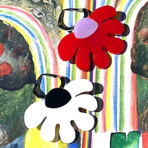 May include: Two flower-shaped earrings. One earring is red with a light purple center, and the other is white with a black center. Both earrings have a silver-colored, geometric loop for attachment. The background features a colorful, abstract design.