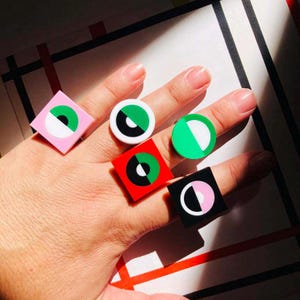 Puede incluir: Cinco anillos coloridos con diseños geométricos. Cada anillo presenta una forma cuadrada o redonda con un diseño de círculo blanco y negro. Los anillos son en tonos de rosa, verde, rojo y blanco.