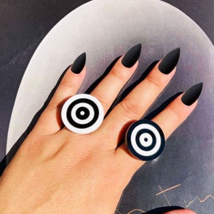 Puede incluir: Dos anillos de blanco y negro con forma de diana, puestos en el índice y el dedo medio de una mano con esmalte de uñas negro.