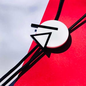 May include: A modern bolo tie featuring a white circular pendant with black geometric shapes, including a triangle and lines. The pendant is set against a red and white background, with black cords extending from the pendant.