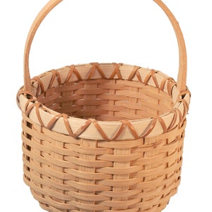 May include: A small, round, woven basket with a natural wood handle. The basket is made of light brown and tan colored wicker.