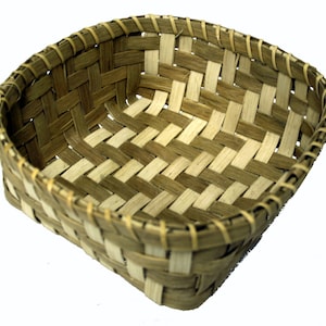 May include: A woven basket made of natural materials, with a light brown and white pattern. The basket is square shaped and has a slightly rounded bottom.