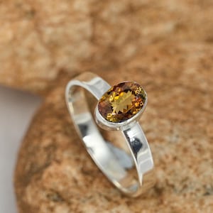 May include: A silver ring featuring an oval-cut gemstone with shades of yellow and brown. The gemstone is set in a simple, polished silver band. The ring is displayed on a textured, brown surface.