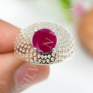 May include: A silver ring featuring a round, deep red gemstone set in the centre. The band has a wide, domed surface with a repeating grid-like pattern of small, square-shaped cutouts across the entire exterior of the metal band.