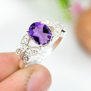 Peut inclure: Une bague en argent avec une améthyste violette centrale de taille ovale. L'anneau est orné de motifs en forme de feuilles de chaque côté, chacun serti de petites pierres blanches rondes et claires qui contrastent avec l'améthyste.