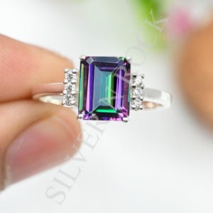 May include: A silver ring featuring a large, rectangular, emerald-cut mystic topaz gemstone with iridescent purple and green hues. The centre stone is flanked by three small, clear, round-cut accent stones on each side of the polished silver band.