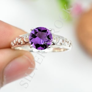 May include: A silver ring featuring a round, faceted purple amethyst gemstone set in a four-prong setting. The band is crafted with an openwork scroll pattern along the sides. The text SILVER ROCK is visible across the centre of the image.