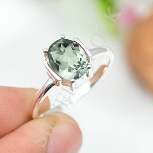 May include: A silver ring featuring a faceted oval-shaped light green gemstone held in a four-prong setting. The band is polished silver, and the stone displays a clear, translucent quality with multiple geometric facets reflecting light.