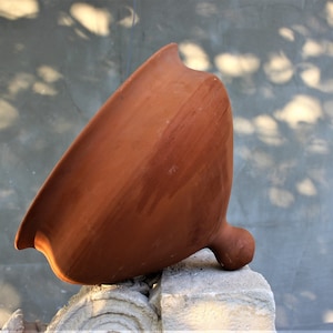 Puede incluir: Una olla de barro grande de color terracota con una abertura ancha y redondeada y una pequeña asa redondeada. La olla está apoyada sobre una superficie de piedra.