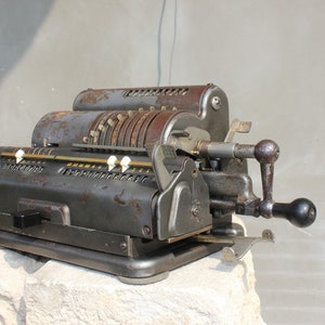 May include: An antique mechanical calculator with a dark gray, weathered finish. The calculator features multiple number dials, white buttons, and a hand crank. The metal shows signs of rust, giving it a vintage appearance. It sits on a light-colored stone.