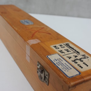 May include: A rectangular wooden box with a metal clasp and labels. The box is light brown with a visible red 'X' mark. Labels indicate measurements of 30 cm. The box appears to be for storage or transport.