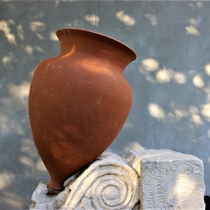 May include: A terracotta-coloured ceramic vase with a rounded body and a wide opening, resting on a white stone base with intricate carvings.