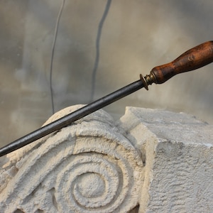 Può includere: Un affilacoltelli in acciaio vintage con manico in legno e un anello in ottone. L'affilacoltelli è appoggiato su una superficie in pietra.