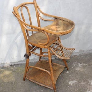 Peut inclure: Une chaise haute ancienne en bois avec un dossier incurvé et un plateau. La chaise a une étagère inférieure et un porte-revues. Le bois a une finition naturelle, montrant des signes d'âge et d'utilisation. Le design de la chaise rappelle les meubles du début du XXe siècle.