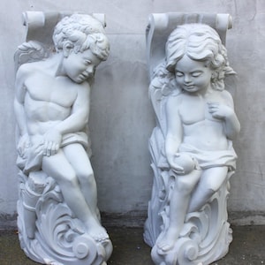 Può includere: Due statue di cherubini in gesso bianco con ali, ciascuna tiene un rotolo e siede su una base decorativa.