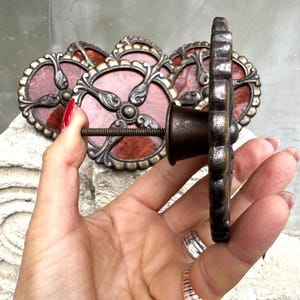 May include: Ornate bronze-toned cabinet knobs with a floral design. Each knob features a circular, reddish-brown center framed by an intricate, decorative border. The knobs are attached with a threaded rod.