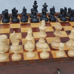 Puede incluir: Un tablero de ajedrez de madera con casillas negras y blancas, y piezas de ajedrez negras y blancas. Las piezas de ajedrez están dispuestas en posición inicial.