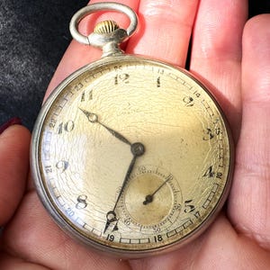 May include: A vintage silver pocket watch with a gold-toned face. The watch features black numerals, a small seconds dial, and black hands. The watch is held in a person's hand.